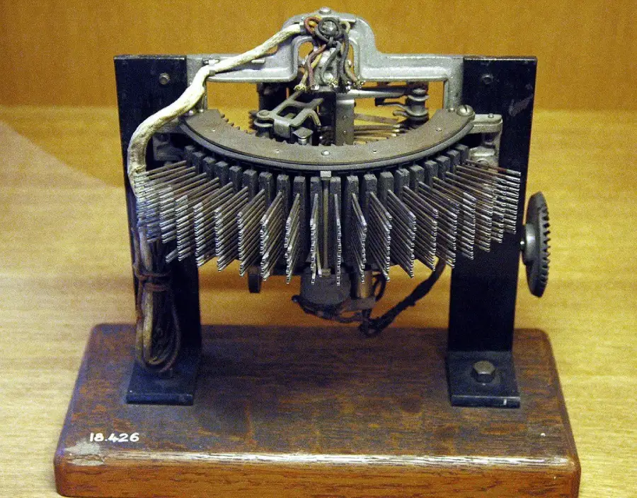 A vintage typewriter mechanism with many metal keys arranged in a semicircle on a wooden base, displaying exposed wires and gears.