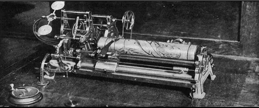 Black and white photo of a mechanical music box with intricate gears, levers, and a large metal cylinder engraved with patterns, sitting on a wooden surface.