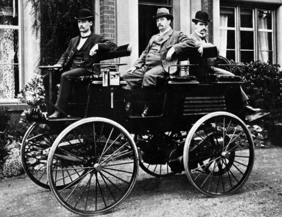 Three men in formal attire and bowler hats sit on an early vintage automobile with large spoked wheels, parked in front of a brick building with tall windows and plants nearby.