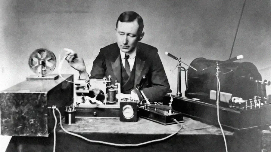 A man in a suit sits at a table with early radio equipment, operating dials and wires, demonstrating historic wireless communication technology. Black and white photograph.