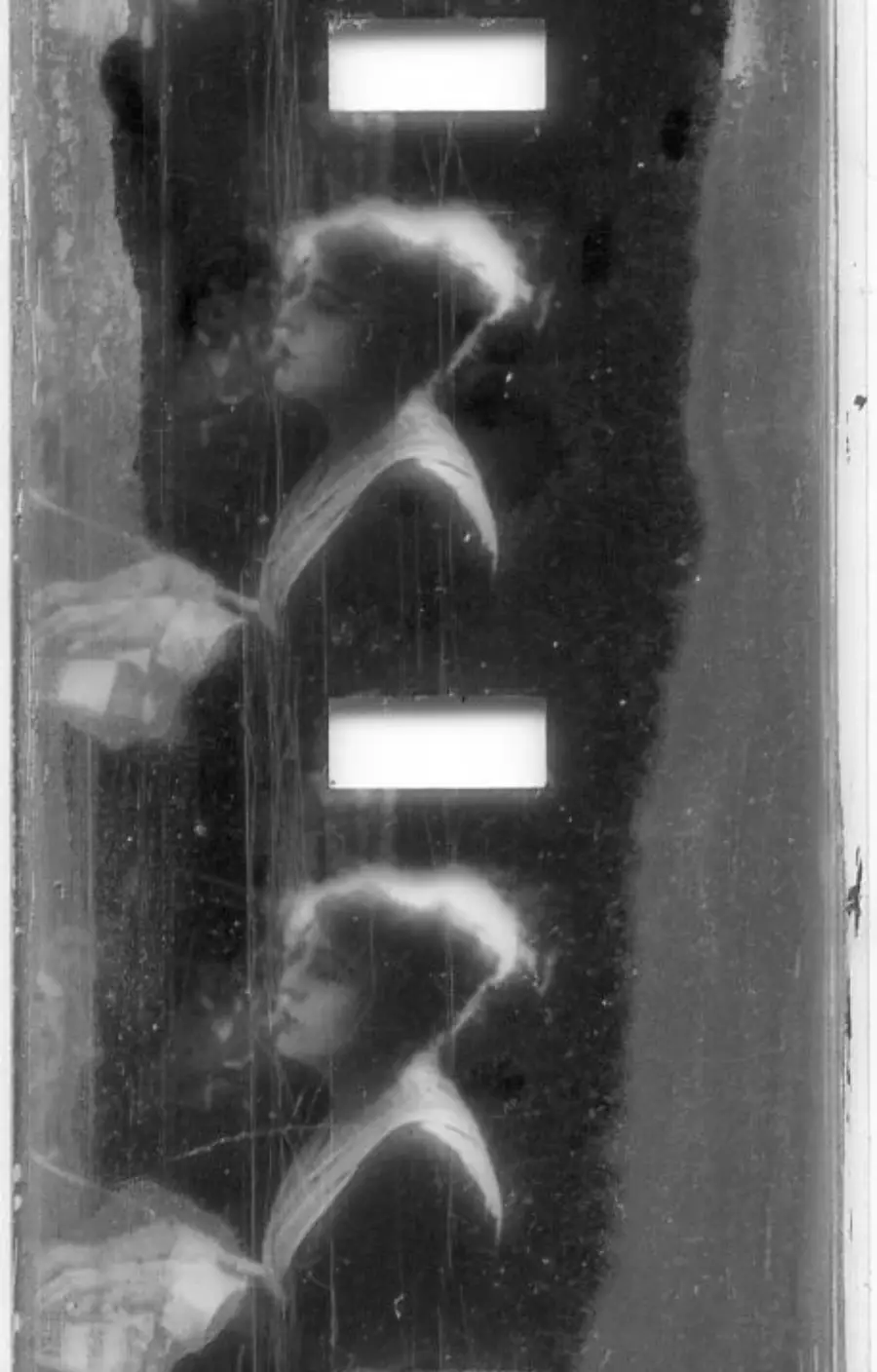 A black and white film strip with two frames showing a woman with glasses and tied-back hair, holding papers and looking to the left. The film strip has visible scratches and rectangular sprocket holes.