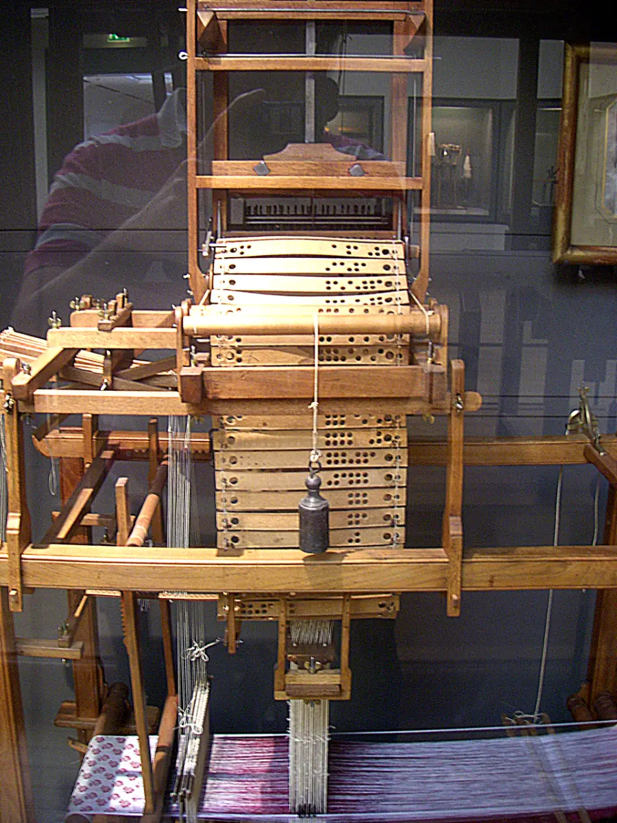 A close-up view of a Jacquard loom, showing its wooden frame and punched cards used to control the weaving pattern. The intricate mechanism and threads are visible in the foreground.