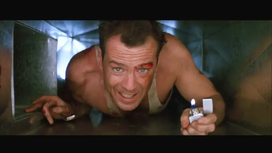 A man with a dirty face and a cut on his forehead crawls through a metal air duct, holding a lit lighter in front of him to see in the darkness.
