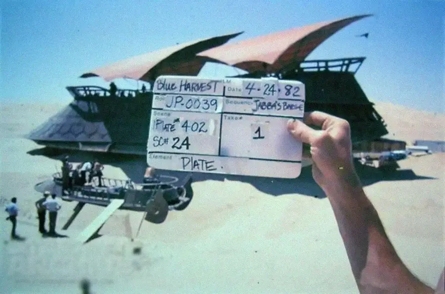 A person holds a movie clapperboard labeled "Blue Harvest" in front of a desert set with large, futuristic vehicles and crew members, during the filming of a scene.