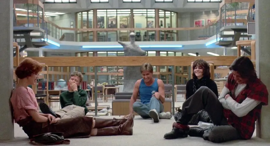 Five teenagers sit on the floor of a spacious library, laughing and talking together. Shelves, railings, and a large statue are visible in the background, creating a warm, relaxed atmosphere.