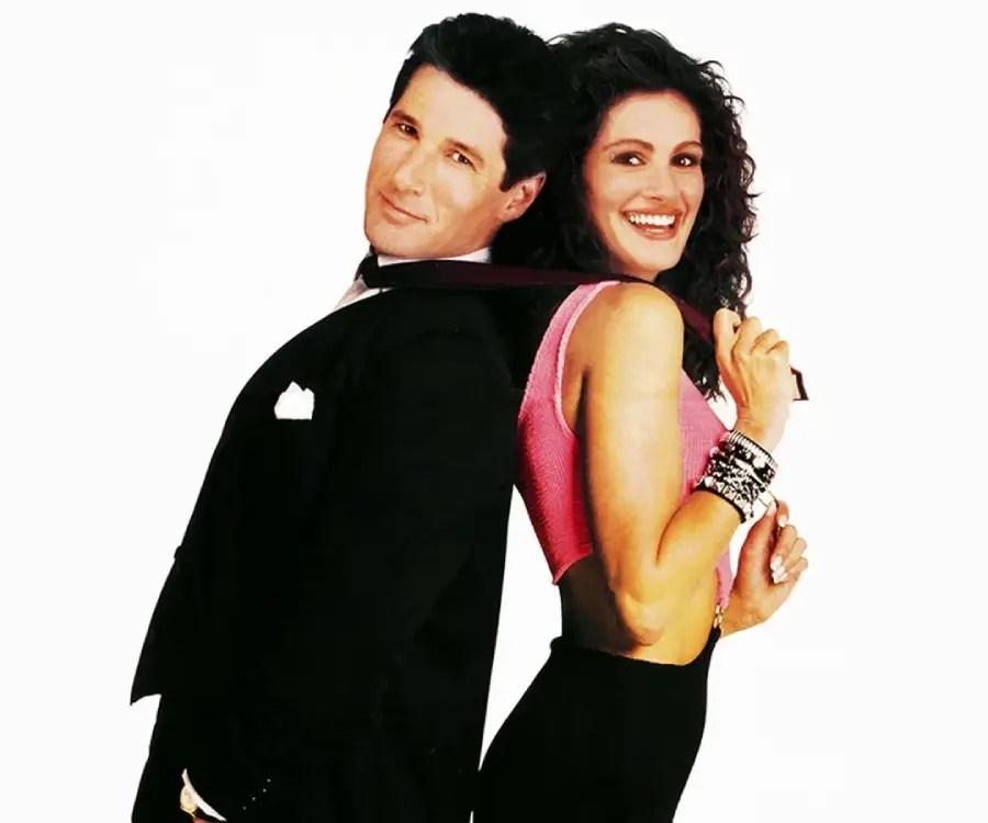 A man in a black tuxedo stands back-to-back with a smiling woman in a pink crop top and black skirt who is playfully holding his tie over her shoulder, both posing against a white background.