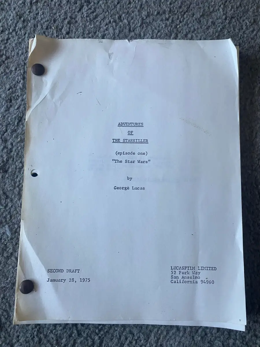 A worn, typewritten script titled "Adventures of the Starkiller (Episode One): The Star Wars" by George Lucas lies on a textured gray surface. It is labeled “Second Draft,” dated January 28, 1975, with company details at the bottom.