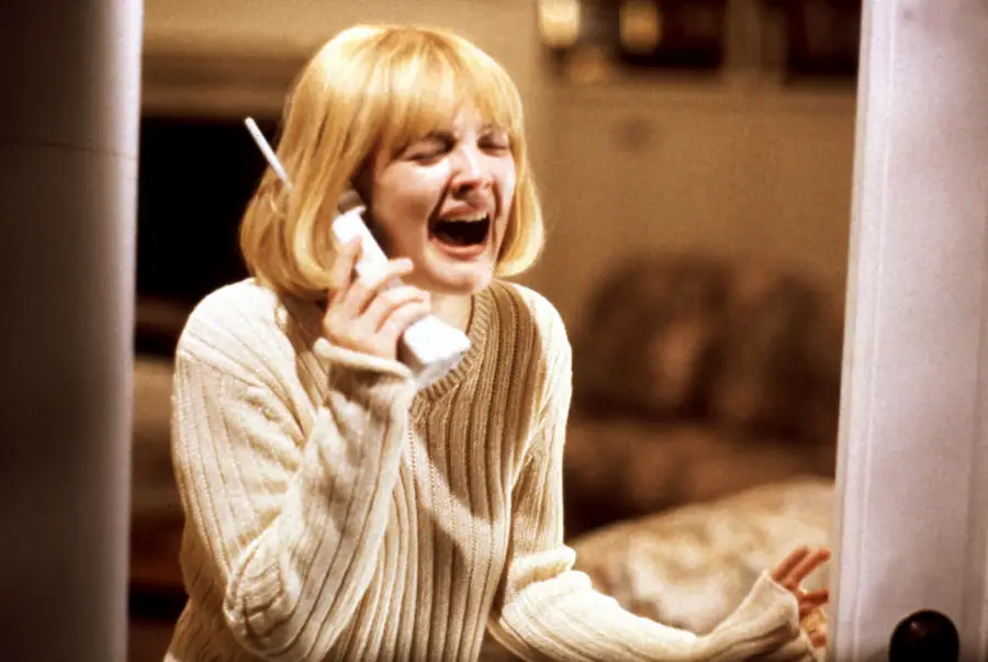 A woman with short blonde hair in a cream sweater holds a cordless phone and screams in fear, standing near a doorway in a dimly lit room.