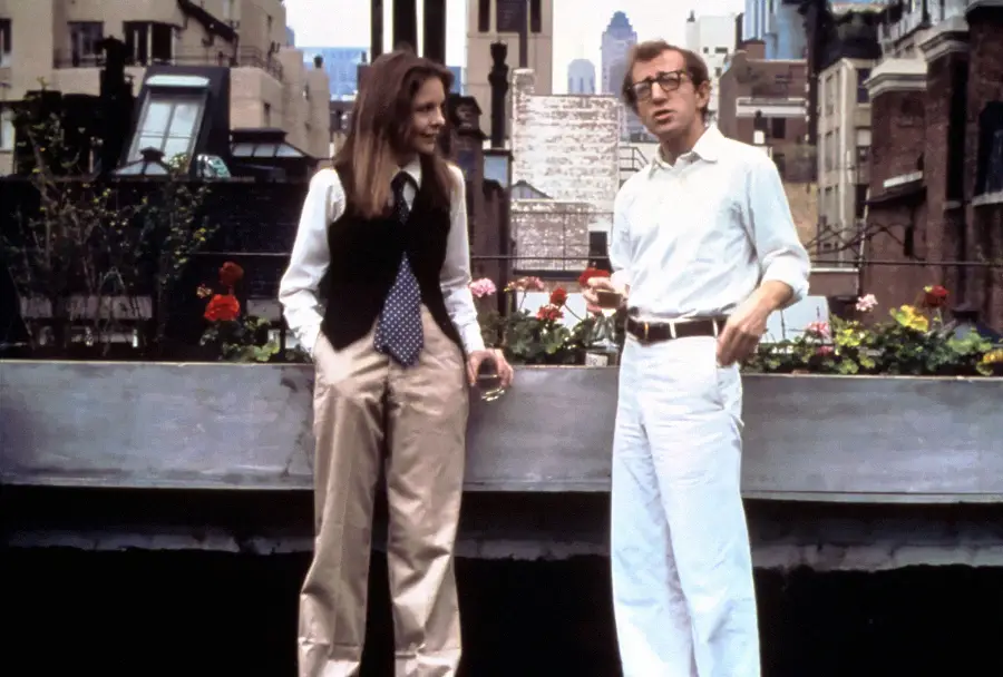 A woman in a vest, tie, and wide-leg pants stands next to a man in white pants and glasses. They are talking on a rooftop with city buildings and flowers in the background.