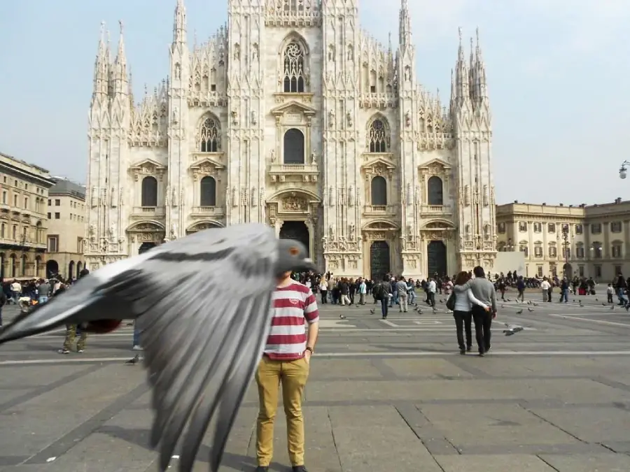 A large pigeon flies close to the camera, partially obscuring a person in a red and white striped shirt standing in front of the Milan Cathedral, with people walking around the plaza.