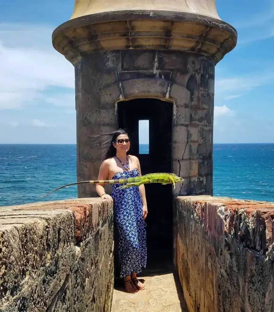 A woman in a blue patterned dress stands by a stone lookout tower near the ocean, smiling as a green iguana rests on the wall beside her under a sunny blue sky.