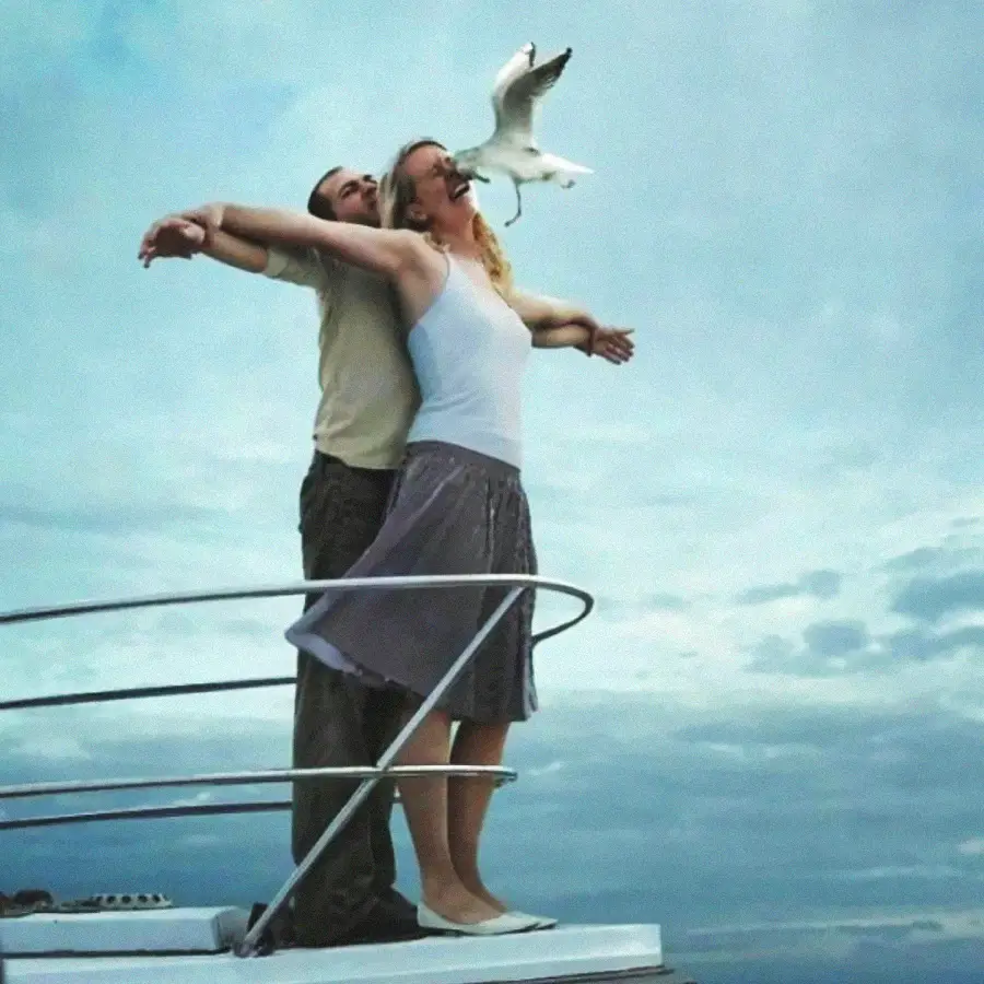 A couple poses at the bow of a boat, recreating the famous Titanic scene with arms outstretched, as a seagull flies by, awkwardly obscuring the woman’s face.