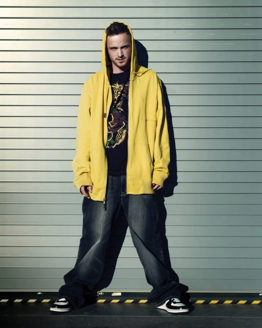 A man wearing a yellow hoodie, black graphic T-shirt, baggy jeans, and sneakers stands against a corrugated metal wall with his hands in his pockets, looking at the camera.