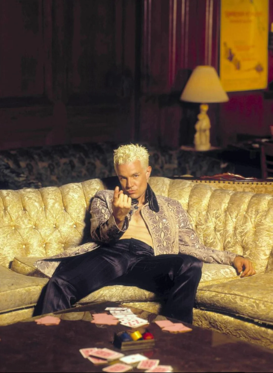 A person with bleached blonde hair sits on a gold ornate sofa, dressed in a shiny patterned jacket and dark pants, gesturing forward with one hand. Playing cards are scattered on the table in front of them.