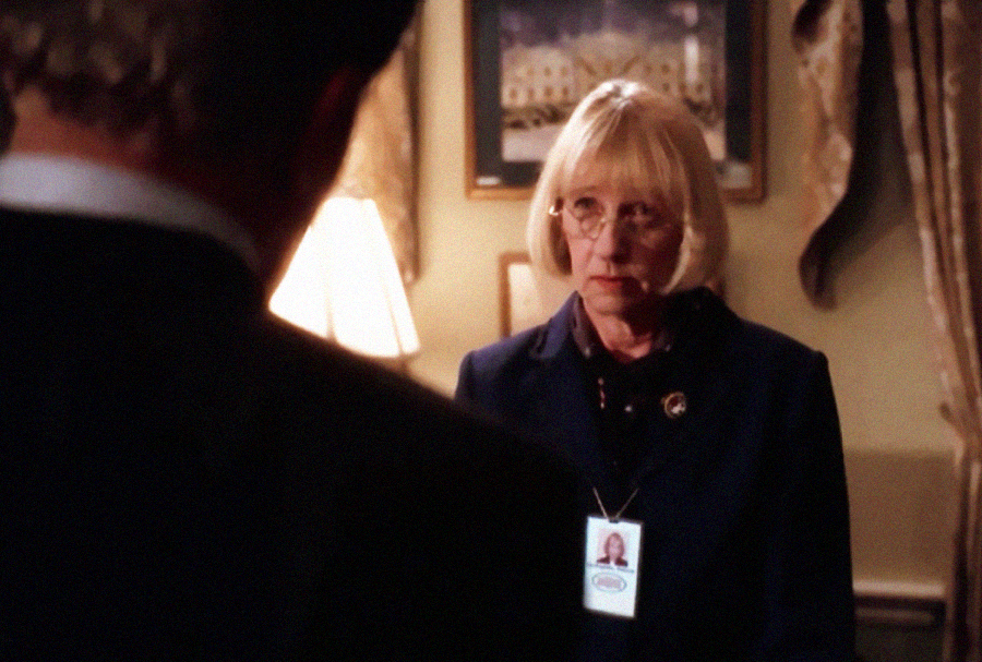 An older woman with blonde hair, glasses, and a visible ID badge stands indoors, facing a man whose back is to the camera. There is a lamp and framed artwork in the softly lit background.