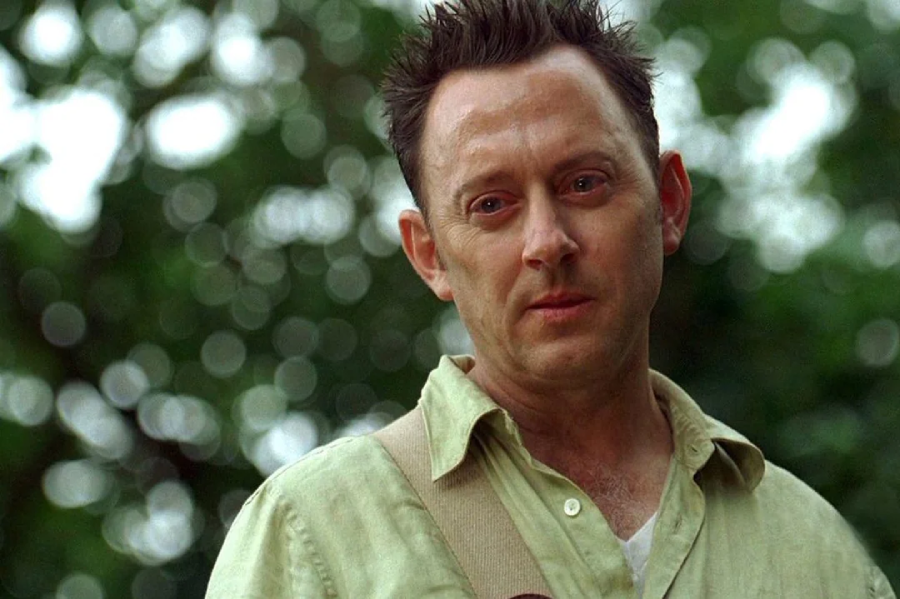 A man with short, spiked hair wearing a light green shirt and a beige shoulder strap stands outdoors, looking slightly downward with a thoughtful or serious expression; blurred green foliage is in the background.