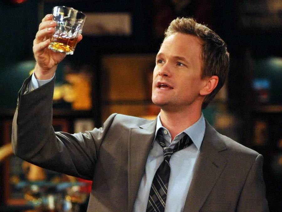 A man in a suit and tie holds up a glass of amber liquid, looking at it intently. The background is a dimly lit bar with blurred details.