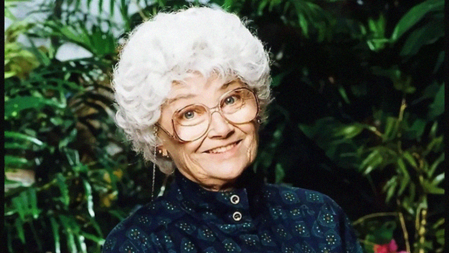 An older woman with short, curly white hair and large glasses smiles at the camera. She is wearing a dark patterned top and is standing in front of green leafy plants.