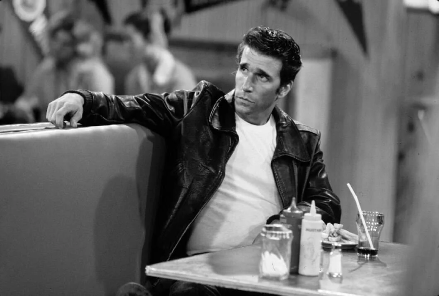A man in a black leather jacket and white T-shirt sits in a diner booth, leaning back with one arm on the seat. There are condiments, a drink, and a straw on the table in front of him. The image is in black and white.