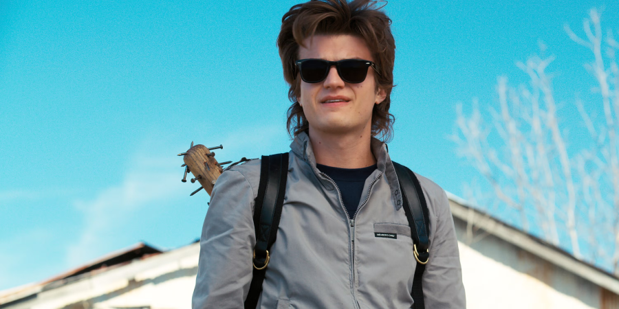 A young man wearing sunglasses and a gray jacket stands outside against a blue sky, carrying a backpack and a spiked baseball bat over his shoulder.