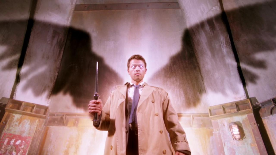 A man in a tan trench coat stands holding a weapon, with glowing eyes and large, dark angel wings cast as a shadow on the wall behind him, in a dramatic, supernatural setting.