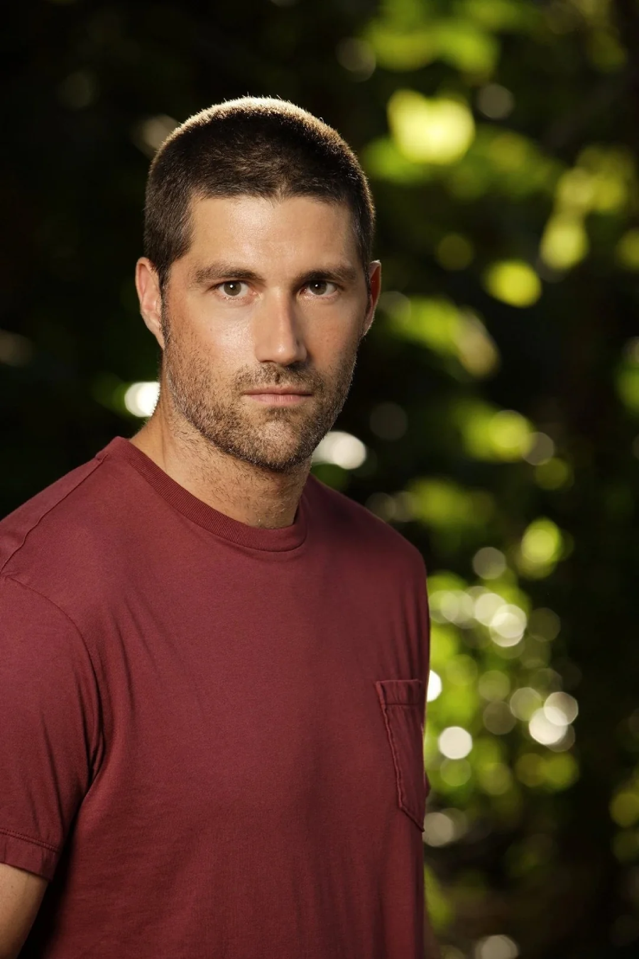 A man with short brown hair and a trimmed beard wearing a maroon t-shirt stands outdoors in front of a background of green foliage and sunlight. He looks seriously at the camera.