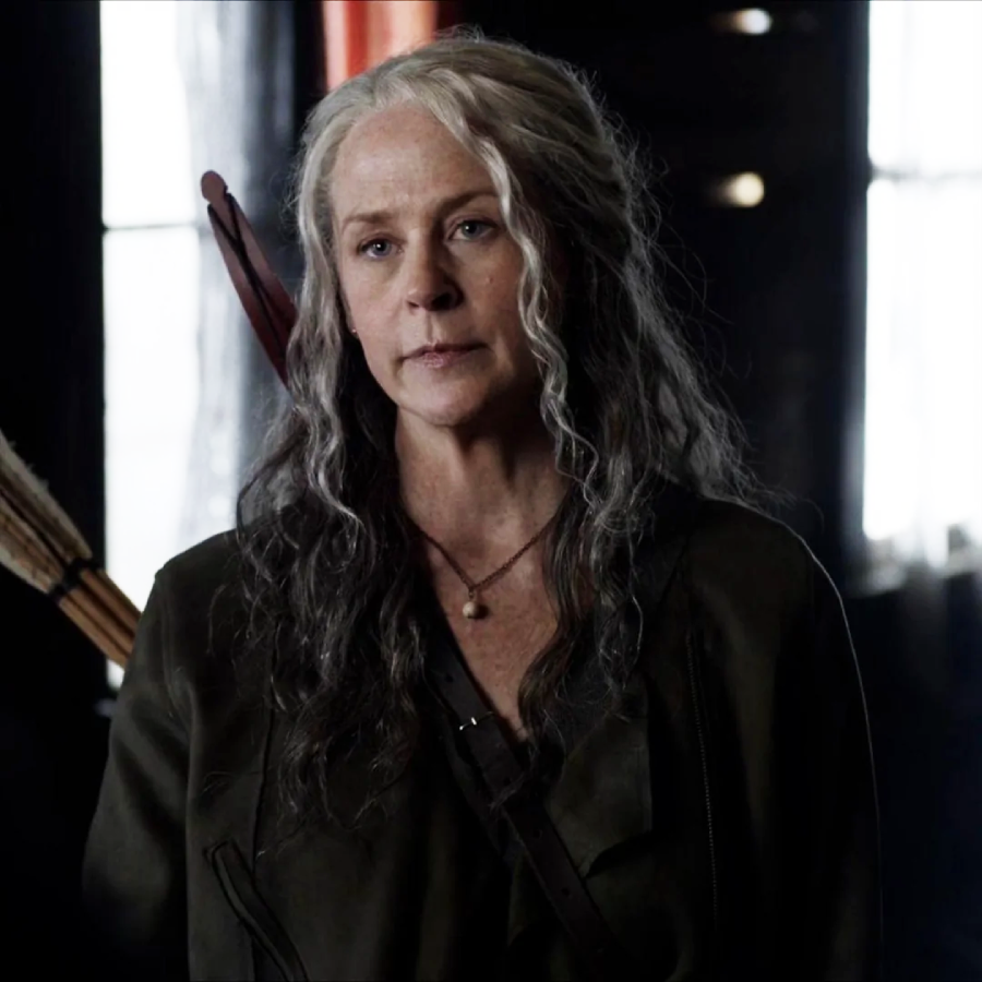 A woman with long, wavy gray hair stands indoors, wearing a dark coat with arrows on her back. She has a serious expression and is lit by natural light from windows behind her.