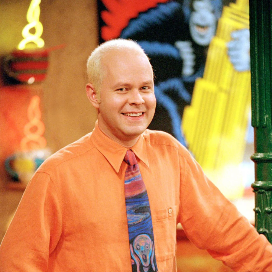 A smiling man with short, bleached blonde hair wears an orange shirt and a colorful tie, standing indoors with vibrant artwork and neon lights in the background.