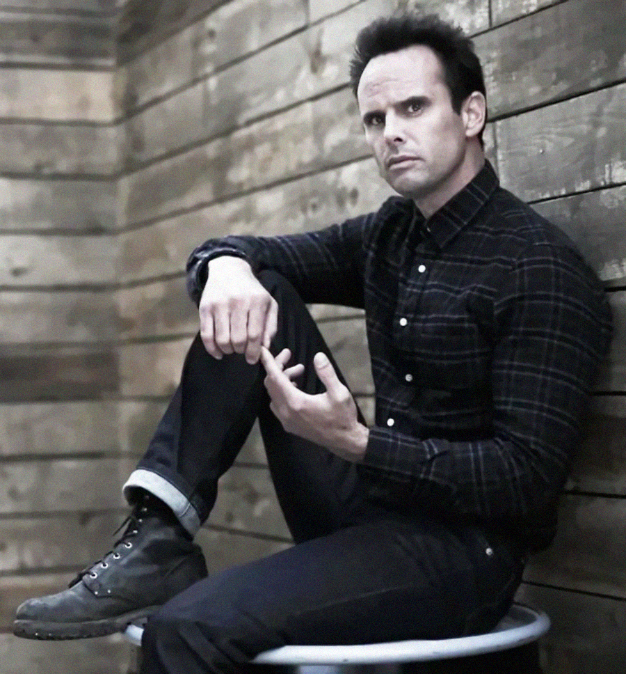 A man wearing a dark plaid shirt, black jeans, and boots sits on a round stool against rustic wooden walls, looking serious with one hand resting on his knee and the other raised in a thoughtful gesture.