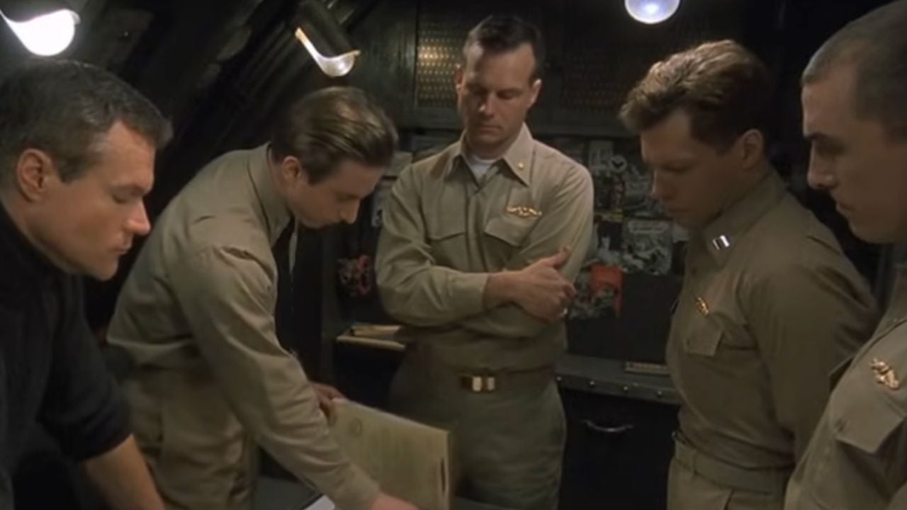 Four men in military uniforms and one in black gather around a table in a dimly lit room, looking at documents, appearing focused and serious.