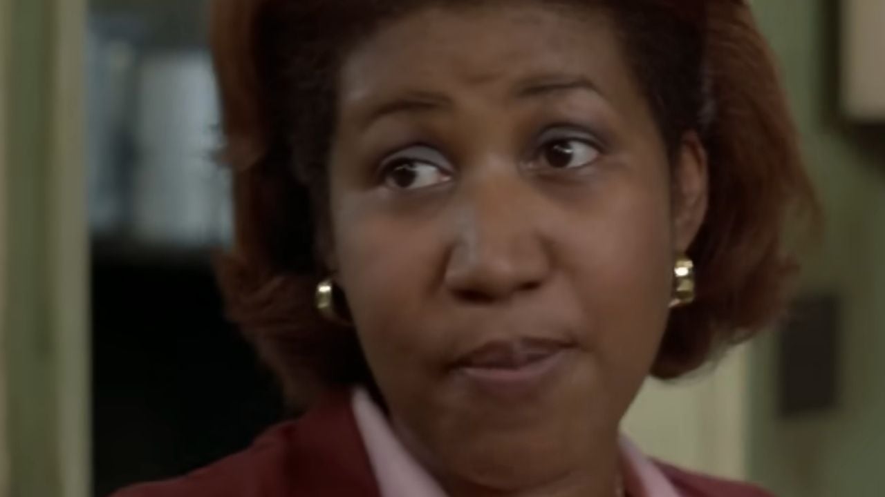 A woman with short brown hair, gold earrings, and a reddish blazer looks to the side with a serious expression. The background is blurry, suggesting an indoor setting.