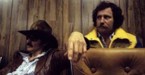 Two men sit side by side in a room with wood-paneled walls. One wears a cowboy hat, sunglasses, and a leather jacket; the other wears a yellow jacket and looks serious. Both are seated on brown chairs.