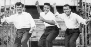 Three men wearing white shirts and dark pants dance energetically in unison, smiling. They are outdoors with metal scaffolding and stage equipment in the background.