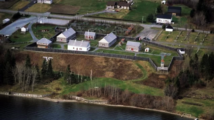 Aerial view of a historic fort with wooden buildings and palisade walls, located near a river. The site includes green lawns, a small tower, and is surrounded by trees and nearby roads and houses.