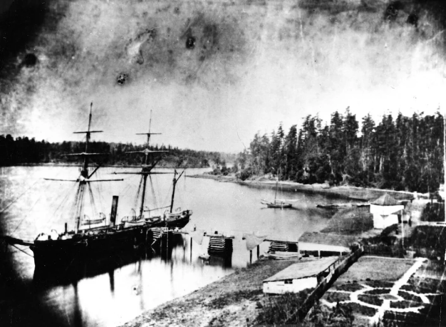 Black-and-white photo of a sailing ship docked at a quiet riverbank, with smaller boats nearby, a forested shoreline in the background, and some wooden docks and pathways in the foreground.