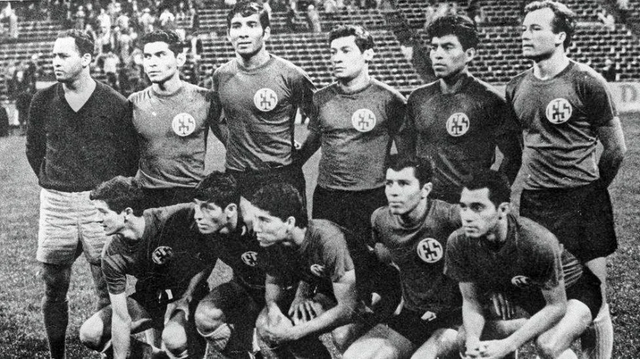 A black-and-white photo of an 11-member soccer team posing together on a field, wearing matching uniforms with a circular emblem on the chest. Spectators are visible in the stadium seats behind them.