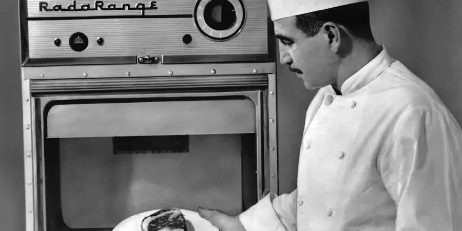 A chef in a white uniform and hat stands beside an early model microwave oven labeled "Radarange," holding a plate of food in front of the open appliance.