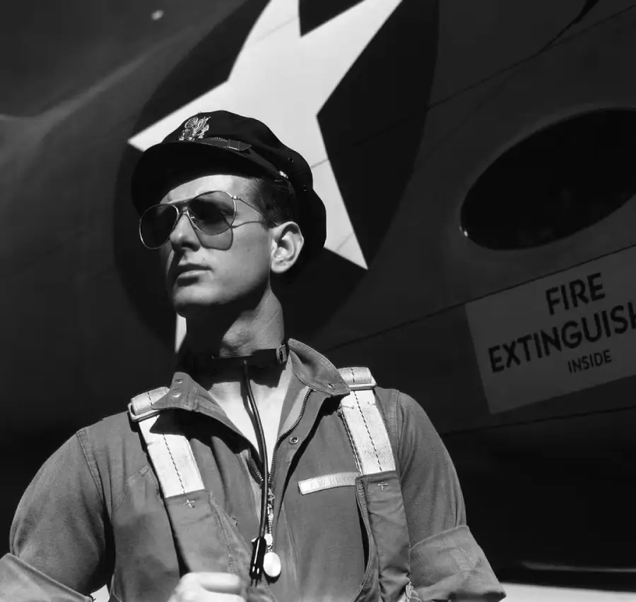 A pilot wearing sunglasses and a cap stands confidently in front of a military aircraft marked with a large white star and a "Fire Extinguisher Inside" sign.