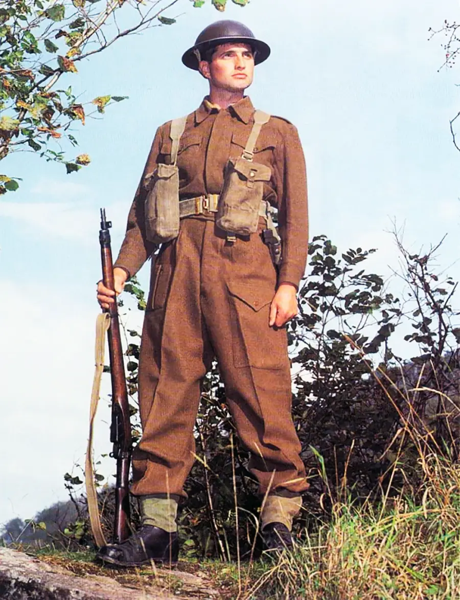 A soldier in brown World War II-era military uniform and helmet stands outdoors holding a rifle. He has gear pouches on his chest, and green foliage is visible in the background.