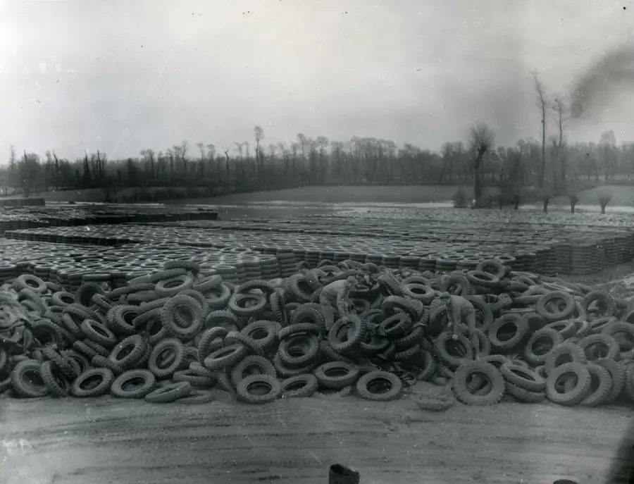 Hundreds of tires are piled in the foreground, with more neatly arranged stacks stretching across a large open field. Leafless trees and a cloudy sky are visible in the background. The image is in black and white.