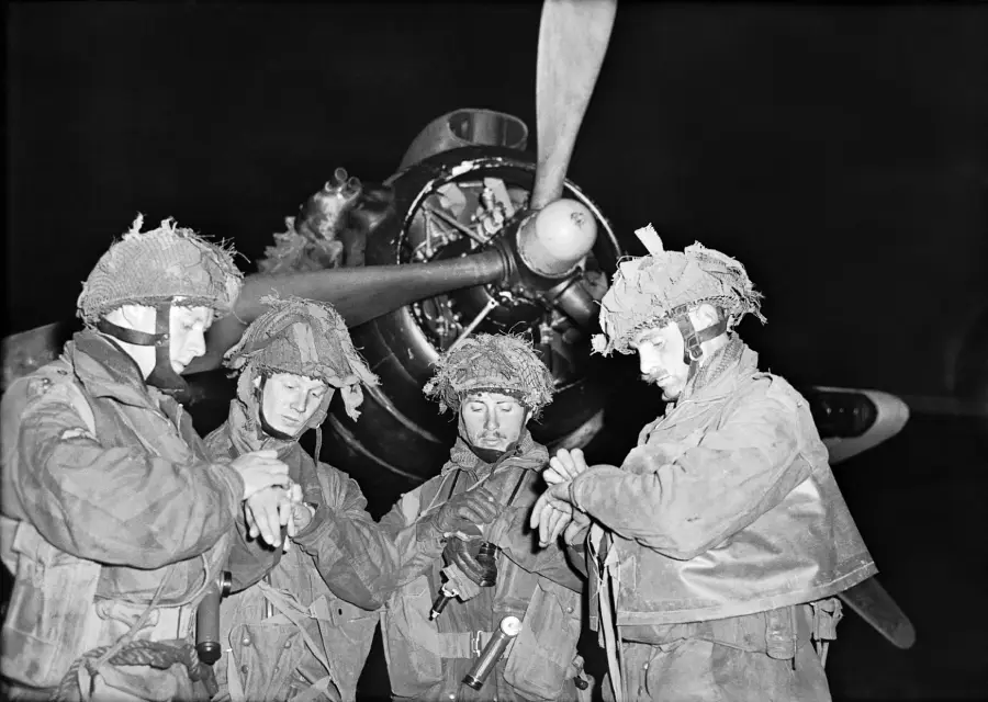 Four soldiers in military gear and helmets stand together at night, checking their watches, with a large aircraft propeller visible in the background. The scene appears intense and focused.