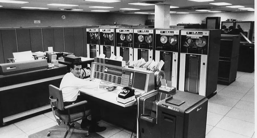 A man sits at a large control console in a vintage computer room with multiple tape drives and mainframe equipment, surrounded by papers, an old telephone, and various office supplies.