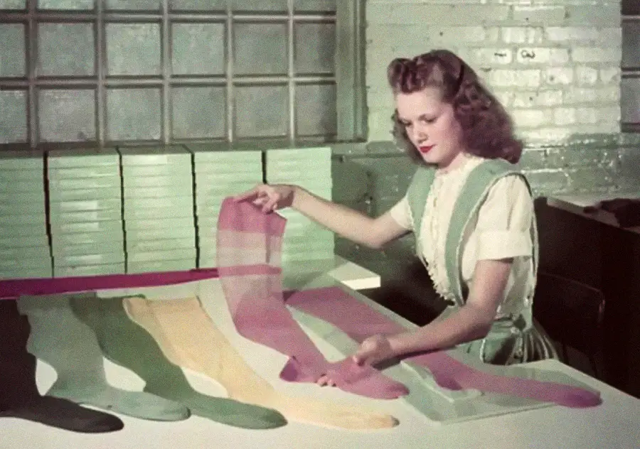 A woman with curled hair sits at a table, inspecting and arranging colorful stockings or nylons in green, yellow, peach, and pink shades, with stacks of green boxes behind her in a vintage setting.