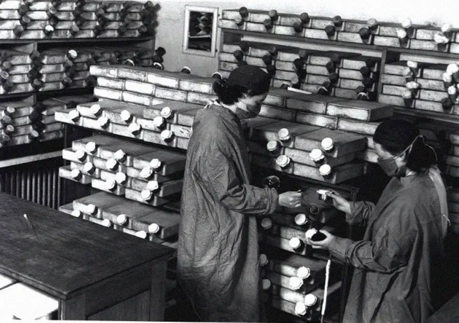 Two women in lab coats and head coverings work with shelves filled with rows of glass bottles or test tubes in a laboratory, possibly conducting experiments or research.