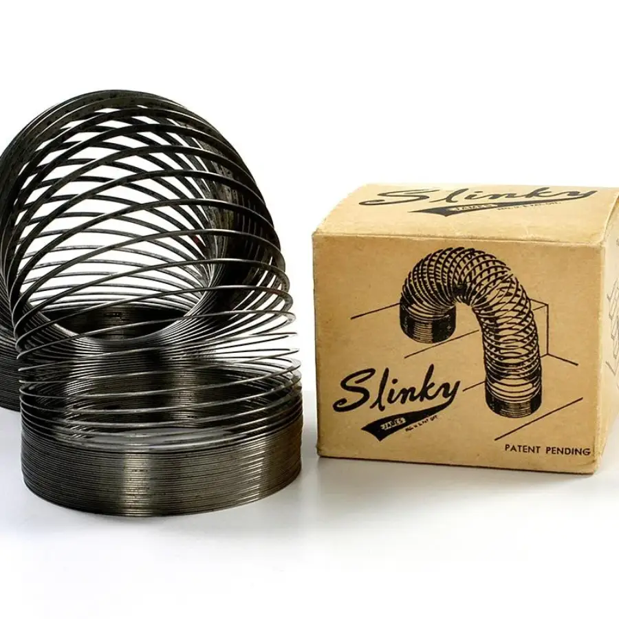 A metal Slinky toy is shown next to its vintage cardboard box with the word "Slinky" and an illustration of the toy on it. The Slinky is partially expanded and standing on a white surface.