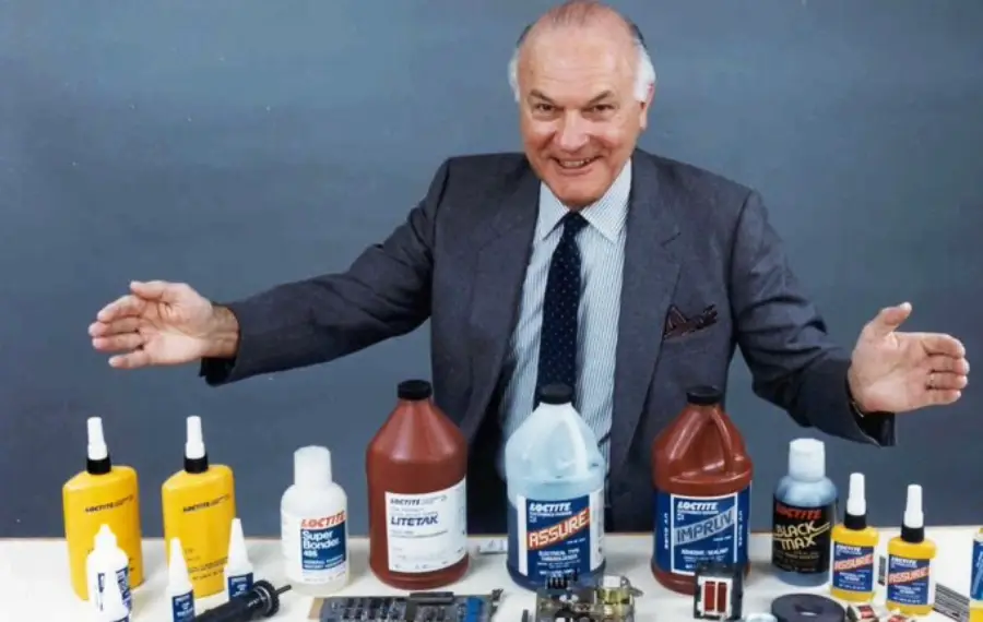 An older man in a suit smiles and gestures with open arms behind a table displaying various bottles and containers of Loctite adhesive products and electronics components against a plain background.