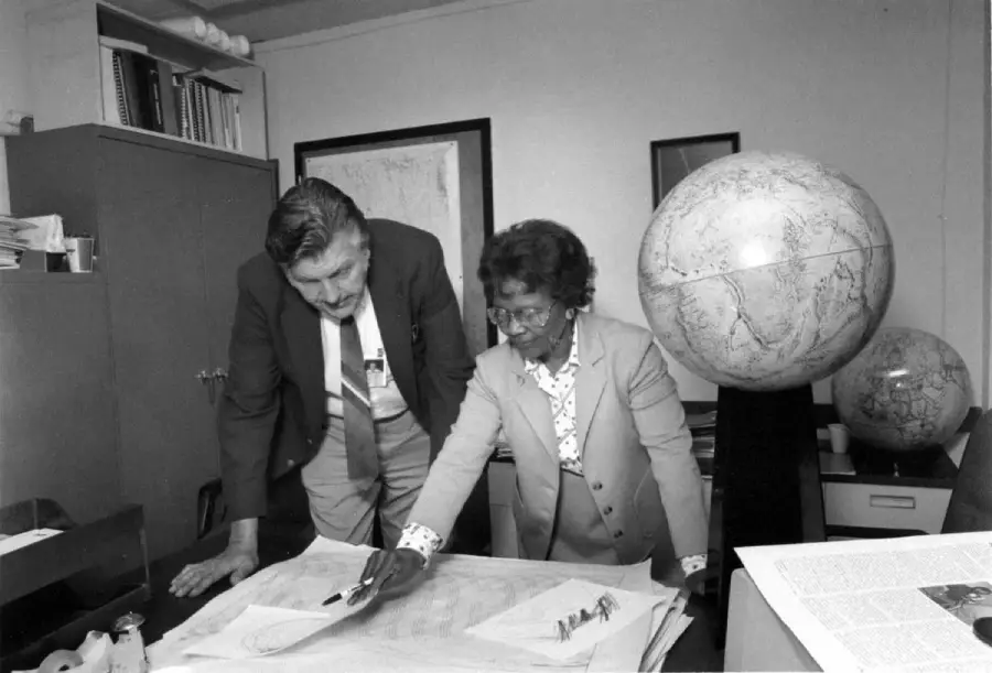 A man and a woman stand in an office, looking at large maps spread on a desk. The woman is pointing at the map. There are two large globes and several bookshelves in the background.