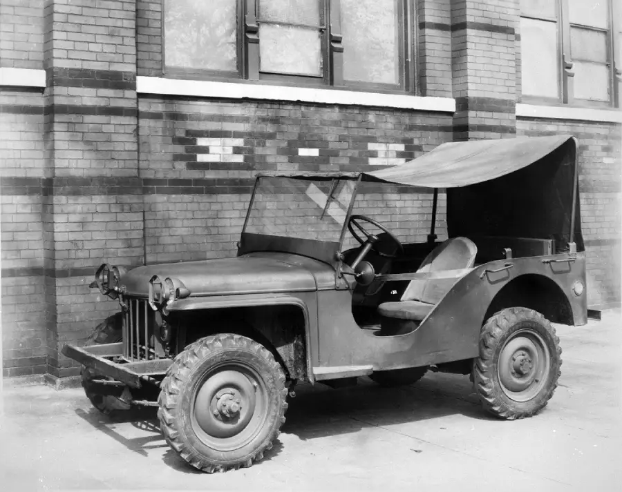 A vintage military-style jeep with a canvas roof is parked on a paved area in front of a brick building with large windows.