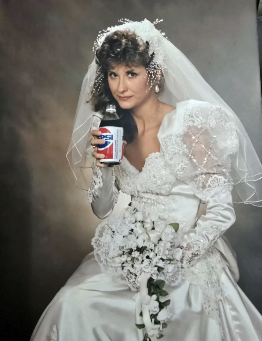 A bride in a white wedding dress and veil sits holding a bouquet of white flowers in one hand and a can of Pepsi in the other, looking at the camera with a slight smile against a studio backdrop.