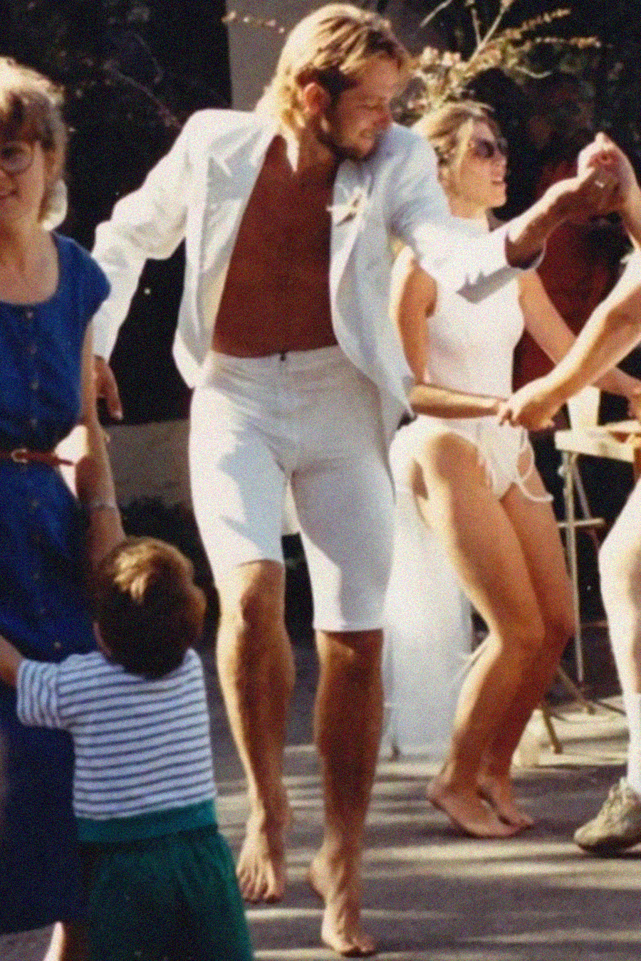 A barefoot man in a white jacket and shorts dances outdoors with a group of people. A woman in blue and a child holding her hand are in the foreground, while other adults in summer clothing join the dance.